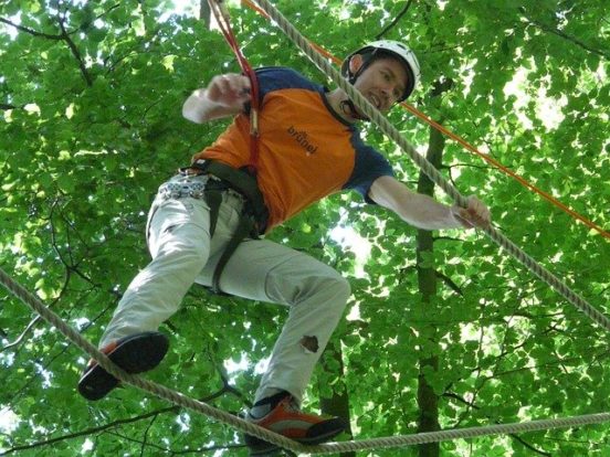 Ein Mann steht auf einem Seil und Klettert. Dabei ist er mit einem Karabiner und einem Helm gesichert. Im Hintergrund sind grüne Blätter zu sehen